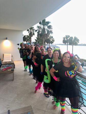 Group of women in neon 1980s-themed outfits and tutus posing on a beachfront resort balcony with palm trees and ocean view, holding inflatable saxophone, boombox and wearing bright leg warmers.