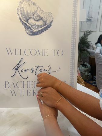 Three hands stacked in front of a bachelorette weekend welcome sign with an illustrated oyster holding an engagement ring — visible engagement and wedding rings, pink manicure and delicate gold bracelets.