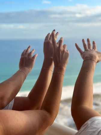 Three sun-kissed arms with delicate bracelets reaching toward a turquoise ocean and blue sky from a beachfront view