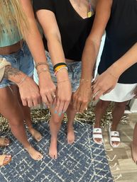 Several friends' arms extended in a casual 'arm party' showing bracelets, rings and blue nail polish; denim shorts, sandals and a blue patterned rug.