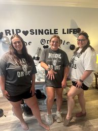 Three smiling women in matching 'Ride or Die' shirts posing at an indoor engagement party with a 'RIP SINGLE LIFE' wall banner, black and silver balloons and celebratory decorations in a living-room setting