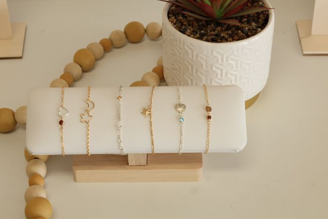 Dainty gold and silver charm bracelets (heart, moon, star, bird) on a white display stand beside a textured white succulent pot and wooden bead garland — minimalist jewelry styling