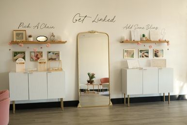 Jewelry boutique interior with gold-framed full-length mirror centered between two white display cabinets, wooden shelves reading “Pick A Chain” and “Add Some Bling,” pink accents and framed ring photos.