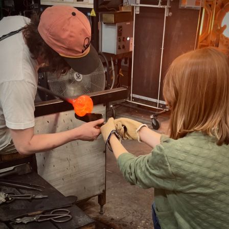 Hands-on glassblowing workshop with an instructor guiding a student as glowing orange molten glass on a blowpipe is shaped while the student holds tools with heat-resistant gloves in a studio.