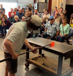 Artisan glassblower shaping glowing orange molten glass on a metal blowpipe during a crowded studio workshop demonstration, audience watching intently.