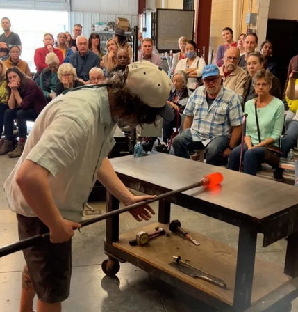 Artisan glassblower shaping glowing orange molten glass on a metal blowpipe during a crowded studio workshop demonstration, audience watching intently.