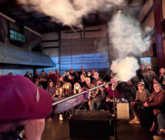 Dim-lit industrial event space where a bundled-up audience watches a performer aim a long fog cannon, sending a dramatic plume of smoke across the room.