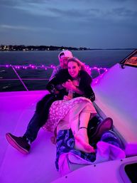 Couple cuddling on a yacht at dusk under purple string lights, woman holding a drink with bare feet and a blanket, calm lake and distant shoreline lights in the background