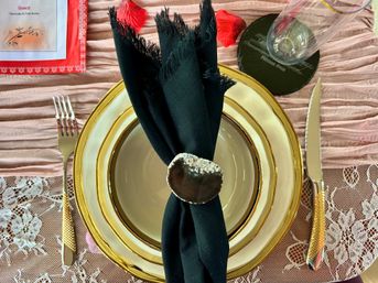 Elegant romantic table setting with gold-rimmed charger and plate, black fringed napkin secured by a chocolate-dipped heart napkin ring, gold-handled cutlery, lace placemat and scattered red rose petals.