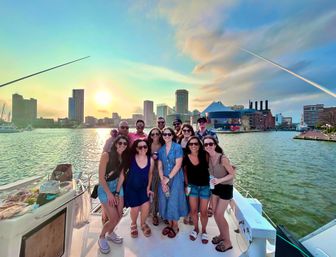 Group of friends on a sunset boat cruise in Baltimore Inner Harbor, smiling with drinks against the city skyline and waterfront