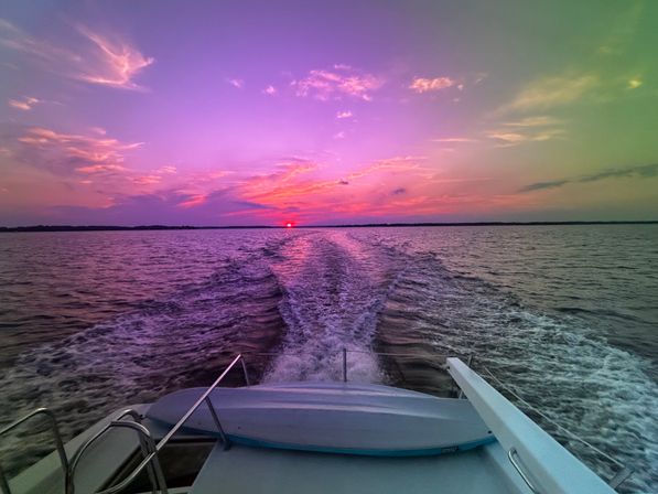 Vibrant pink and purple sunset over open water, view from a boat stern with a small kayak on deck and a foamy wake leading toward the glowing sun on the horizon.