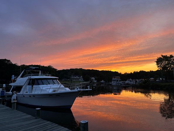 White motor yacht moored at a wooden dock in a quiet marina during a vivid orange and purple sunset, streaked sky reflecting on glassy water with tree-lined shoreline and waterfront homes.