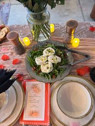 Outdoor reception tablescape with burrata-topped arugula salad and pecans on a decorative platter, gold-rimmed plates, menu card, glass vase of flowers and LED votive candles.