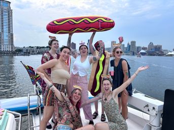 Boat party on an urban harbor: smiling group of women on a boat holding an oversized inflatable hot dog and wearing novelty hot-dog headbands, city skyline and waterfront in the background.
