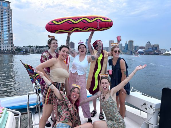 Boat party on an urban harbor: smiling group of women on a boat holding an oversized inflatable hot dog and wearing novelty hot-dog headbands, city skyline and waterfront in the background.