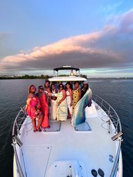 Group of eight women posing on the bow of a white yacht during golden-hour sunset over calm coastal waters, wearing colorful summer dresses with dramatic pink clouds overhead