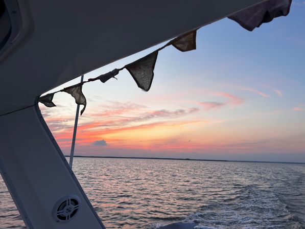 Sunset boat-deck view with pink-orange sky over open water, gentle wake trailing behind and weathered pennant flags strung from the cabin.