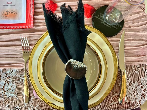 Top-down view of an elegant place setting: gold-rimmed charger and plate, black linen napkin with a white-speckled decorative napkin ring, gold textured flatware, lace placemat and soft pink table runner for a romantic celebration.
