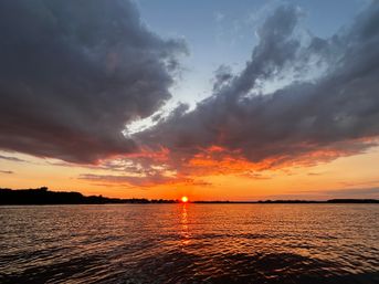 Fiery orange sunset over a calm lake with dramatic dark clouds, sun reflected as a glowing path on rippled water and a silhouetted shoreline on the horizon.