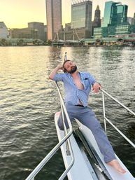 Relaxed man lounging on the bow of a boat, barefoot with an open blue checkered shirt, leaning on the rail while an urban waterfront skyline glows at sunset.