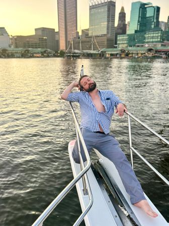 Relaxed man lounging on the bow of a boat, barefoot with an open blue checkered shirt, leaning on the rail while an urban waterfront skyline glows at sunset.