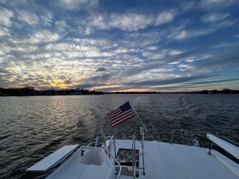 View from a boat stern with an American flag fluttering above the wake on coastal waters at sunset, dramatic layered clouds and a distant shoreline.
