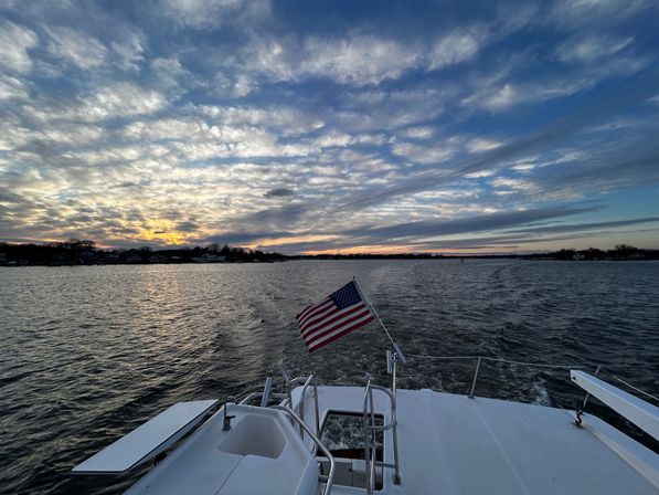 View from a boat stern with an American flag fluttering above the wake on coastal waters at sunset, dramatic layered clouds and a distant shoreline.