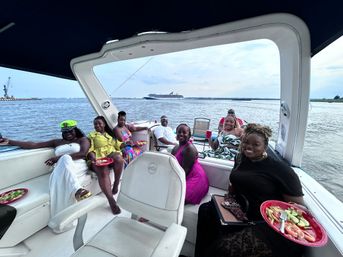 Smiling group enjoying a covered party boat, holding red plates of shrimp and salad while relaxing on the water with a cruise ship and harbor in the distance.
