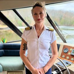 Smiling yacht captain in a white uniform with gold epaulets standing at a boat helm inside the wheelhouse, lake and wooded shoreline visible through angled windows.