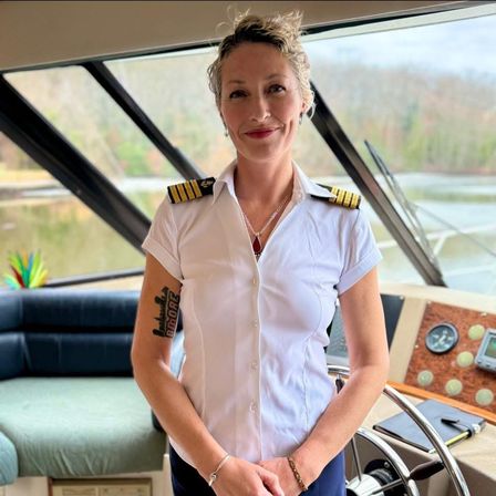 Smiling yacht captain in a white uniform with gold epaulets standing at a boat helm inside the wheelhouse, lake and wooded shoreline visible through angled windows.
