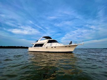 Sleek white motor yacht anchored on calm coastal waters beneath a bright blue sky with wispy clouds and a distant tree-lined shoreline.