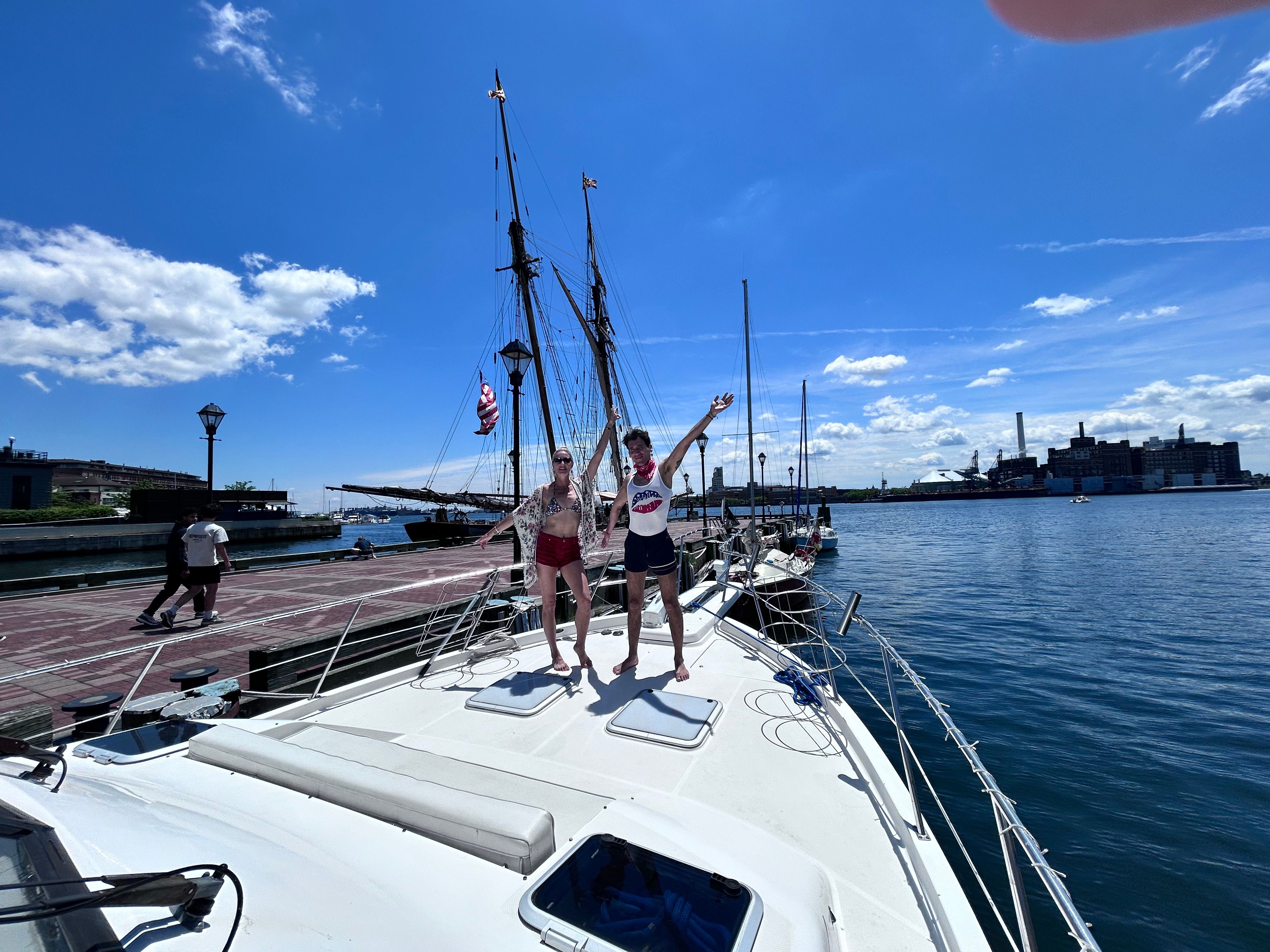 Two people posing on the bow of a white sailboat at a sunny harbor with tall ship masts, an American flag, waterfront docks and a city skyline under a bright blue sky