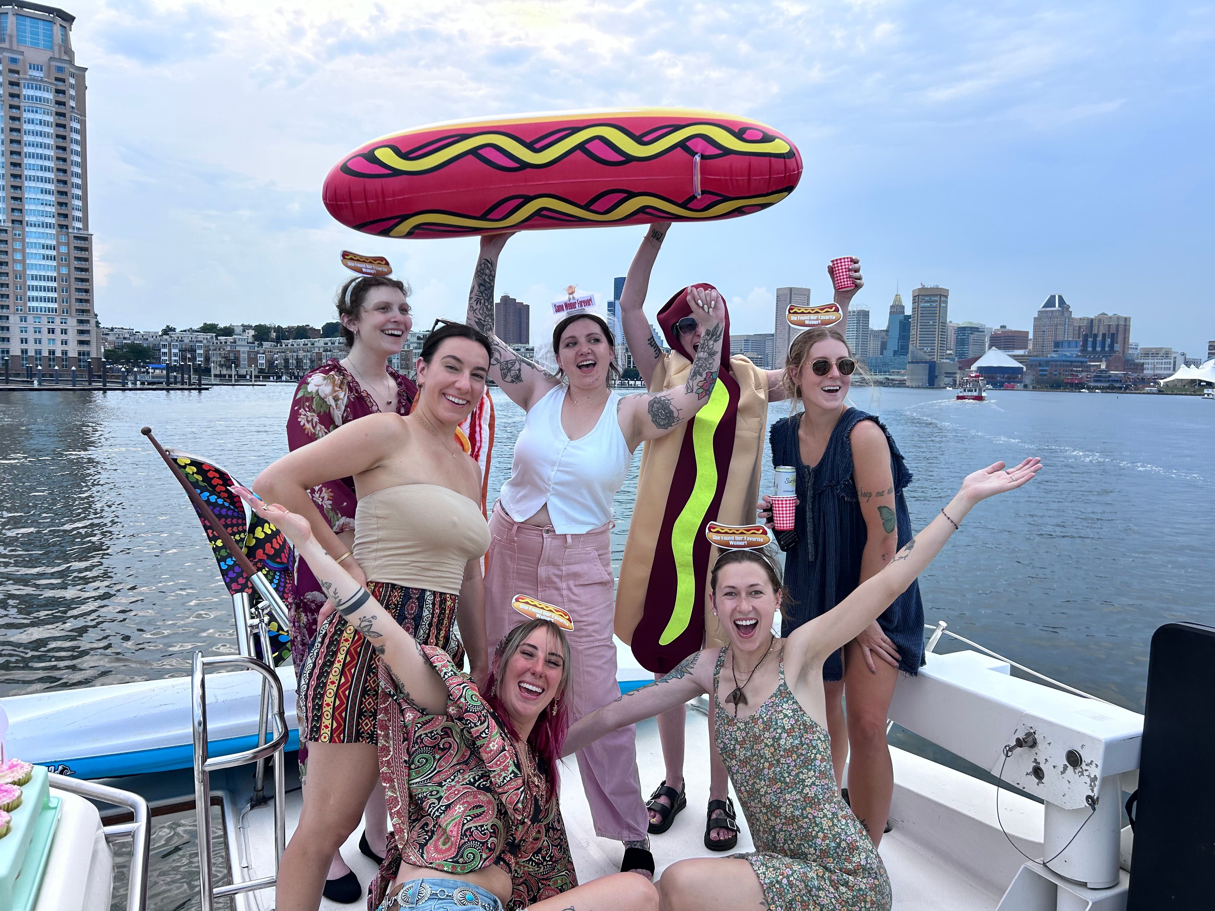 Friends having a summer boat party on a harbor with a city skyline backdrop — one in a hot dog costume holding a giant inflatable hot dog while others cheer with drinks.
