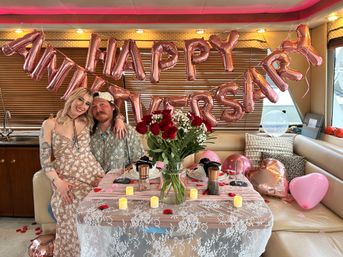 Smiling couple seated in a yacht cabin under rose-gold HAPPY ANNIVERSARY balloons, with red roses, flickering candles, lace-covered table and pink heart balloons for a romantic celebration
