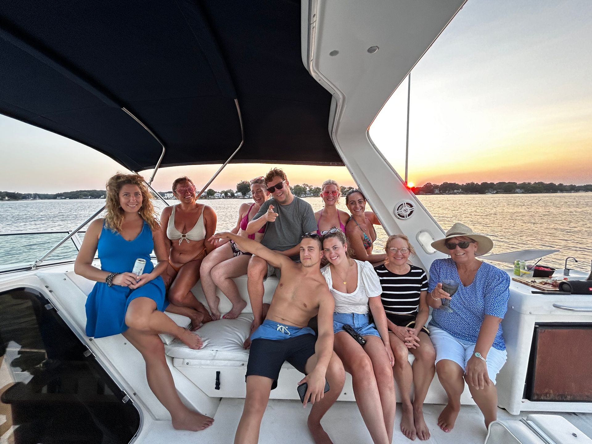 Smiling group of people relaxing on a motorboat at sunset on a calm lake, wearing swimsuits and summer clothes, lounging under a canopy with the shoreline and orange sky in the background.