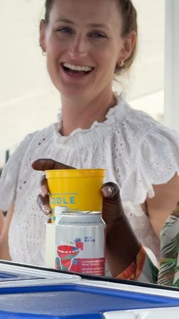 Smiling person in a white eyelet blouse at a sunny outdoor gathering, a hand holds a bright yellow plastic cup above a strawberry-flavored canned cocktail on a blue table.