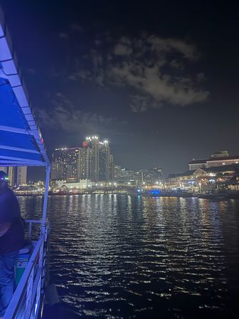 Nighttime waterfront skyline with illuminated high-rise hotels and waterfront restaurants, boats docked and colorful lights reflecting on rippling harbor water; boat deck with blue awning in foreground.