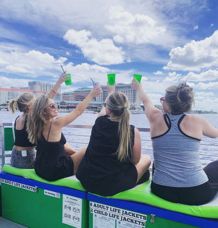 Four friends seated on green life-jacket boxes raise neon cups in a joyful cheers on a sunny boat ride along a city waterfront with skyline and fluffy clouds overhead.