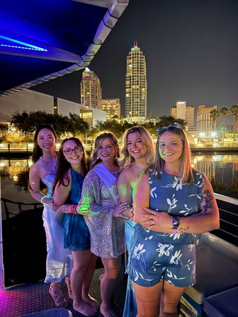 Five smiling women in party outfits on a lit waterfront boat at night, a bride-to-be wearing a sash, with downtown high-rise skyline and reflections on the water in the background.