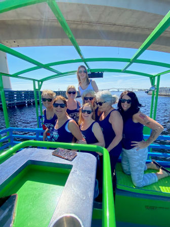 Smiling group of women in matching navy tank tops and sunglasses posing on a bright green open-air party boat under a bridge, with blue sky and waterfront in the background.