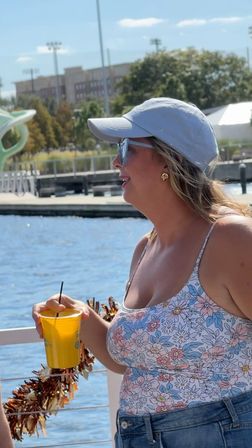Person in a light baseball cap and sunglasses wearing a floral tank top and denim shorts, holding a yellow cup with a straw on a sunny marina dock by blue water and waterfront buildings, festive garland on the railing.