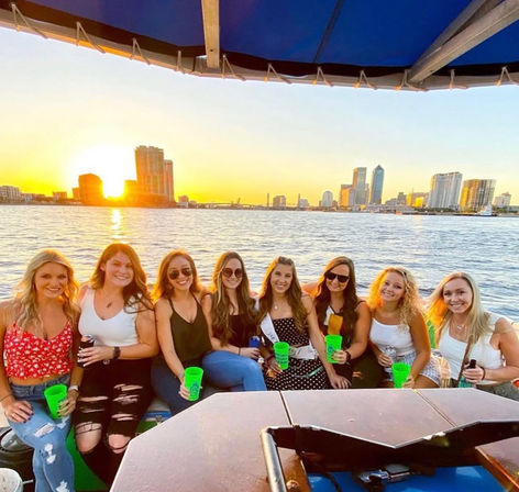 Group of eight friends on a sunset river cruise, smiling and holding bright neon cups with a downtown waterfront skyline behind them.