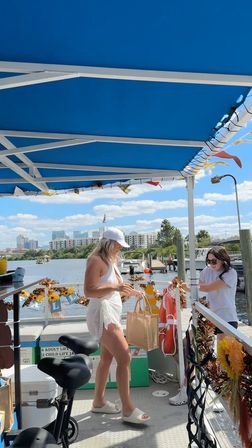 Sunny waterfront scene: two people on a decorated boat deck under a blue canopy exchanging a paper bag at a marina with docked boats and a city skyline in the background.