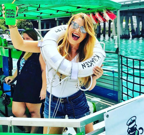 Smiling woman in sunglasses on a bright green pedal boat with a life preserver around her shoulders, holding up a green cup that says 'PEDAL PEDAL DRINK!' with turquoise water and a pier in the background.