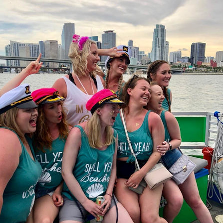 Laughing bachelorette group wearing captain hats and matching teal 'Shell Yeah' tank tops on a party boat with Miami skyline and bay in the background.
