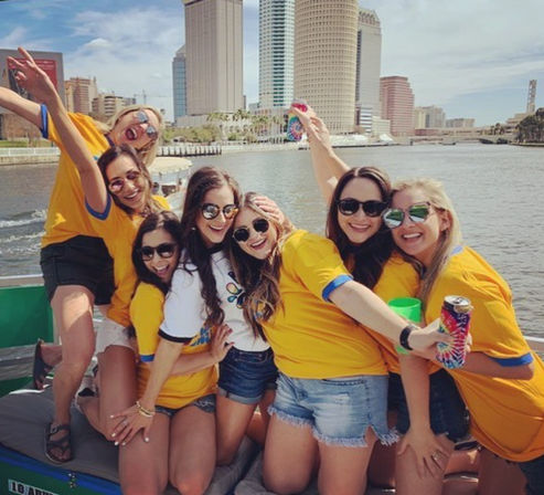 Cheerful group of friends in yellow shirts and sunglasses celebrating on a sunny city riverboat with high-rise skyline in the background.