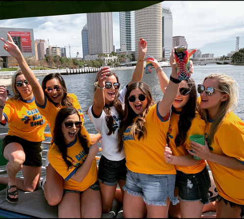 Group of smiling women in matching yellow t-shirts cheering with drinks on a boat in front of a sunny downtown waterfront skyline.