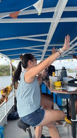 Woman in sunglasses and denim shorts raising her arms at a covered party-boat bar with drinks, shoreline and water visible