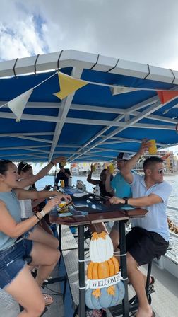Friends toasting with yellow cups at a bar on a covered party boat under a blue canopy, festive pennant flags and waterfront buildings visible