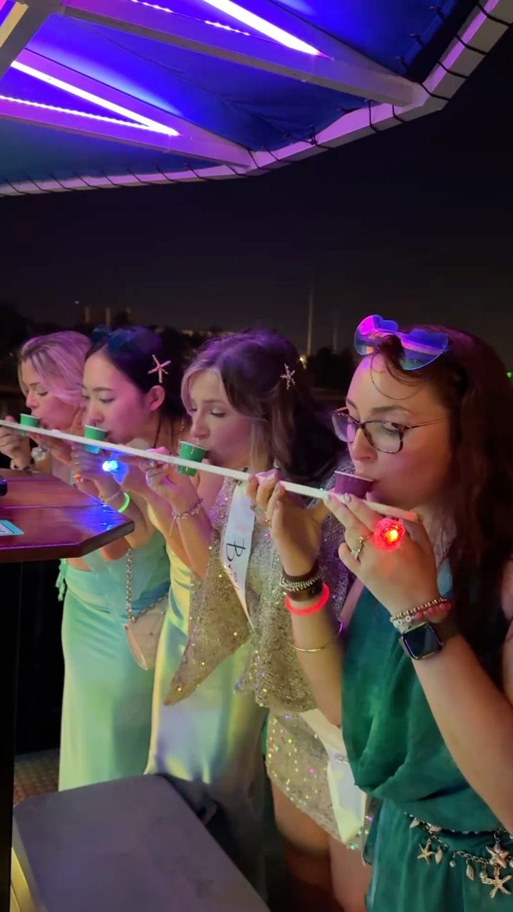 Four women in party dresses taking shots from a long shot-ski under neon lights at an outdoor bar at night, wearing glow bracelets, star hair clips and a bachelorette sash.